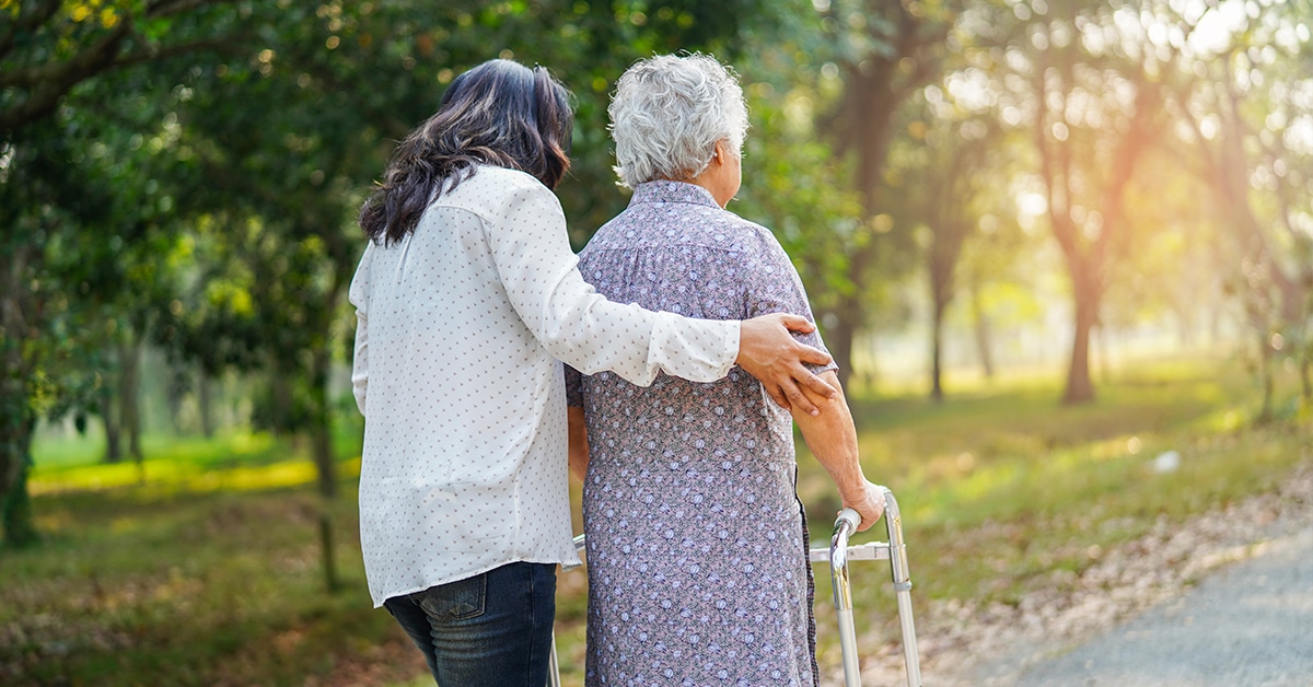 A caregiver supporting an older woman with a cane while walking in a park illustrates the importance of expert Medicaid planning.