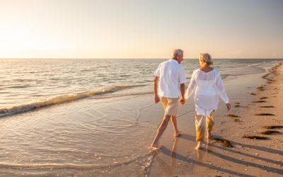 An Elderly Couple In White Shirts And Khaki Pants Walk Along The Water On A Beach At Sunset.