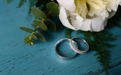 Two Silver Wedding Bands On A Teal Blue Table Next To A White Carnation Flower With Green Leaves.