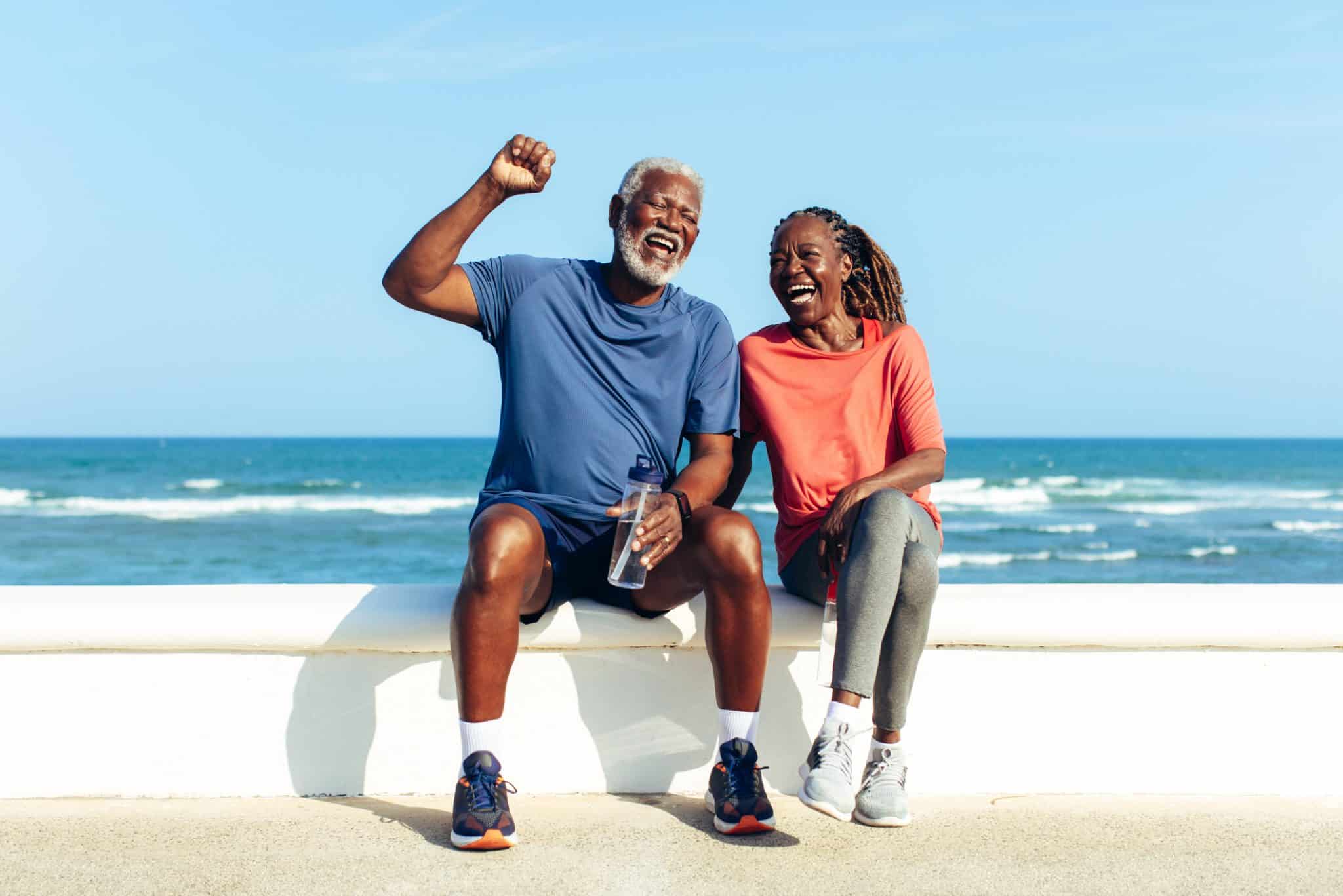 An older husband and wife feeling strong after exercise near a sunny beach; they represent healthy aging.