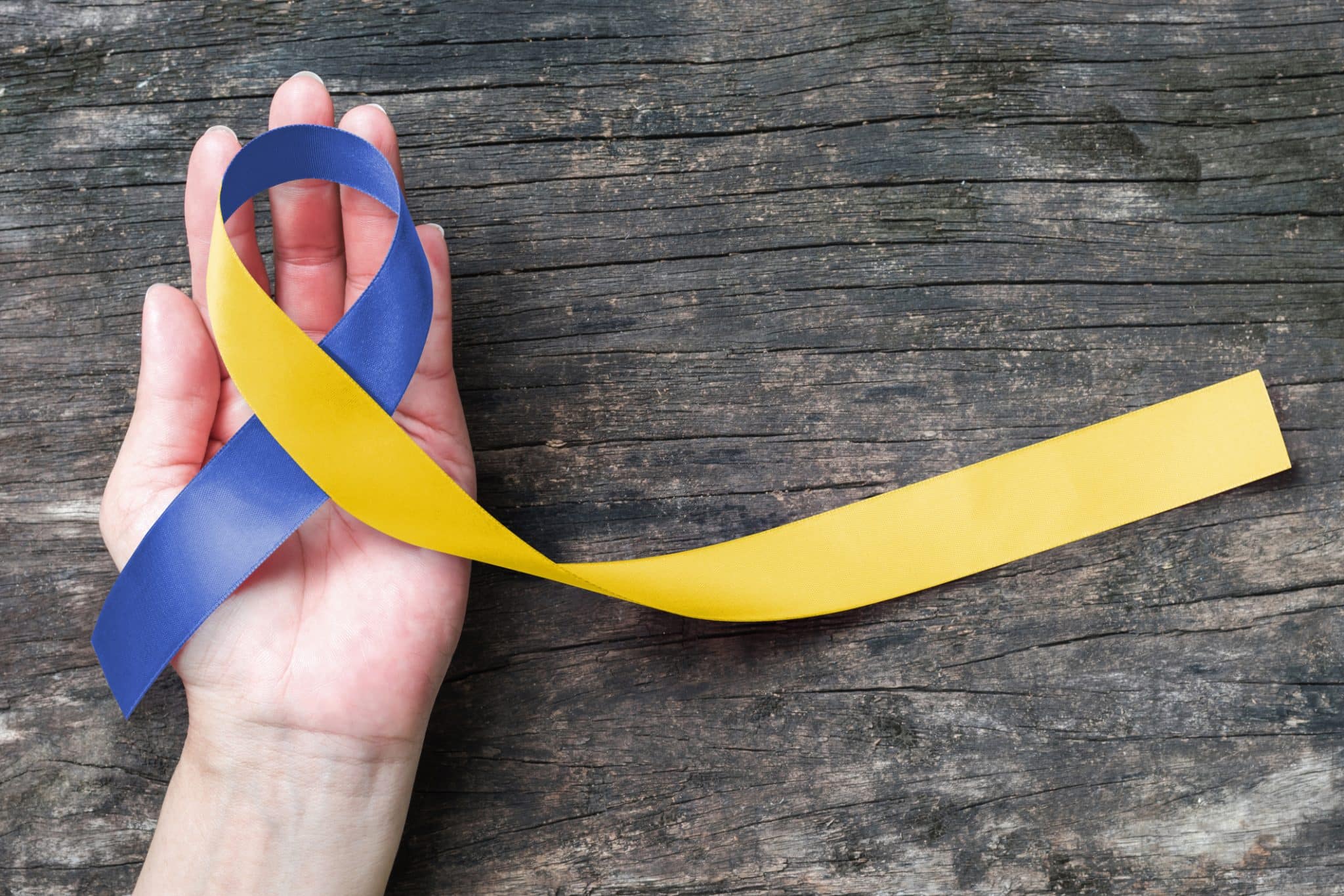 A hand holding a blue and yellow ribbon with the Down Syndrome Awareness Month colors on a wooden table.