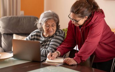 An older woman and her adult daughter sit together at a kitchen table reviewing documents related to Medicaid planning.