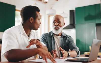 An adult son sits at a kitchen table reviewing paperwork with his elderly father.