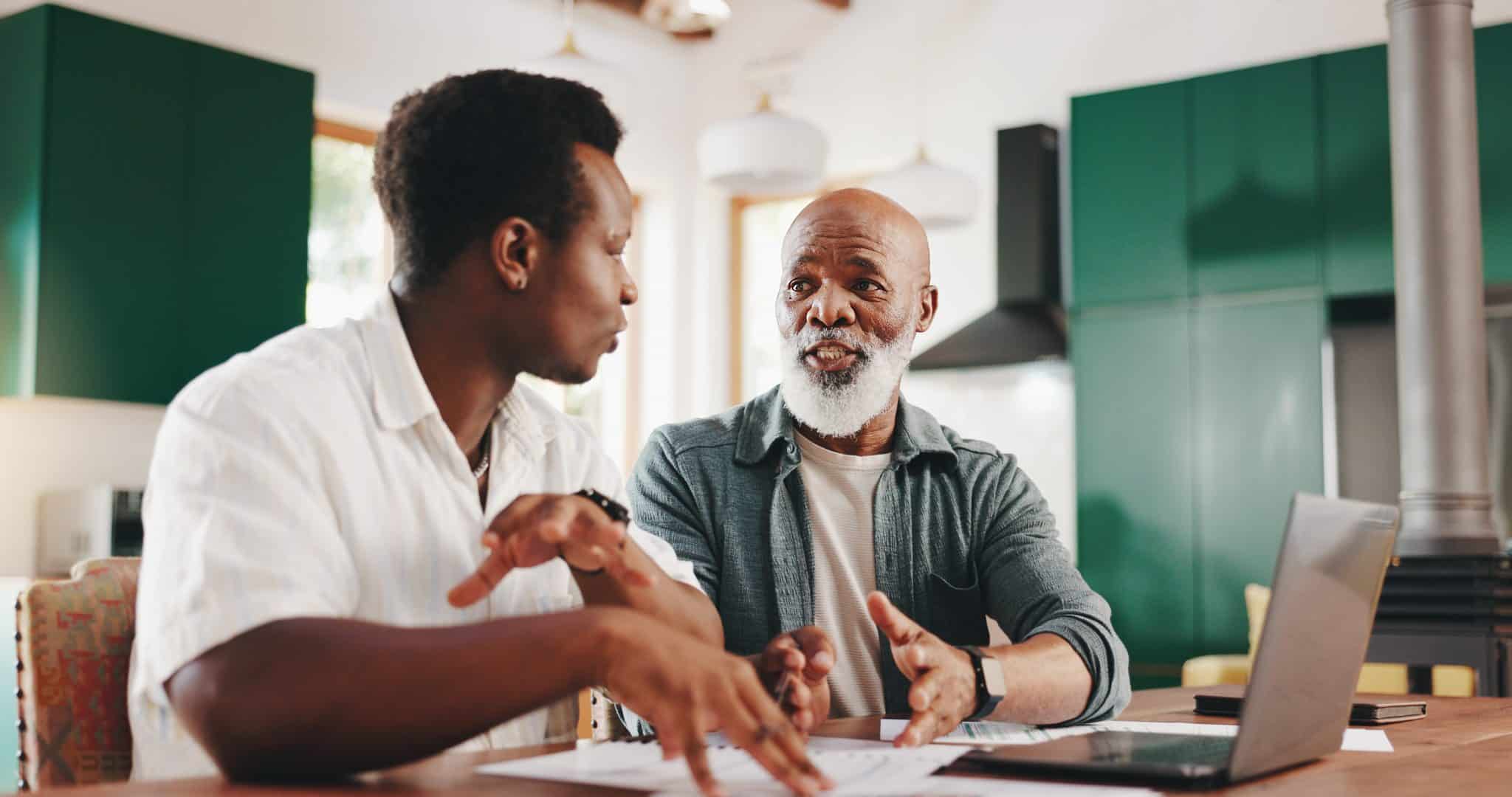 An adult son sits at a kitchen table reviewing paperwork with his elderly father.