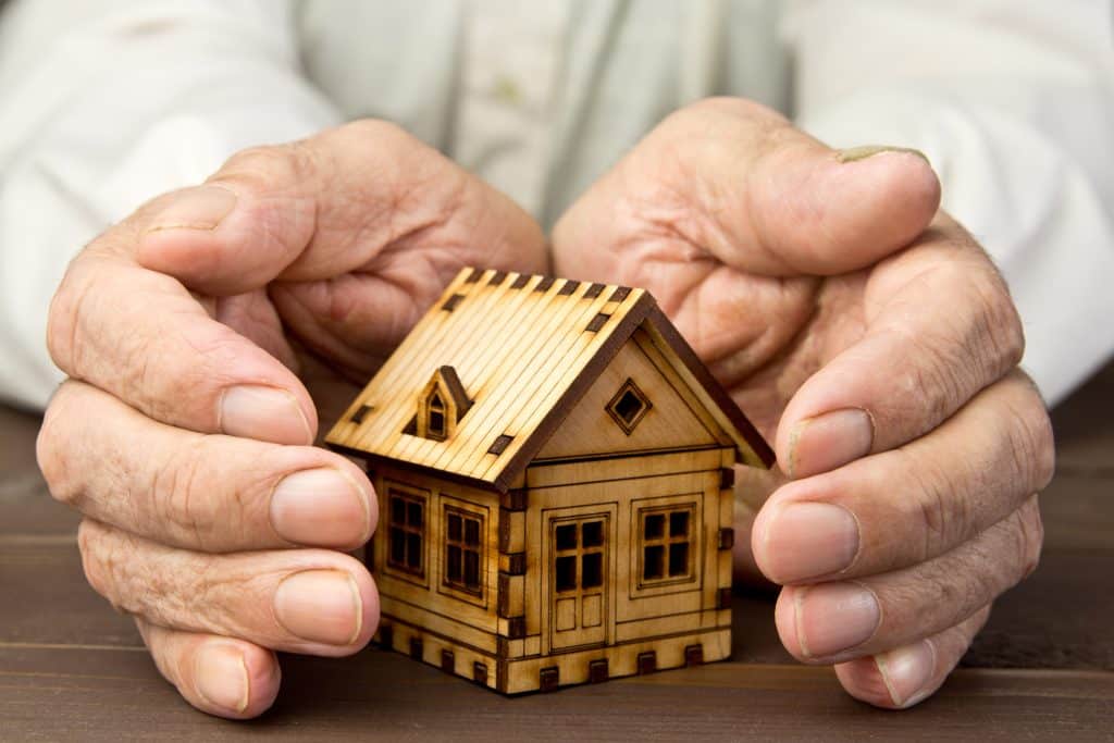 : A senior man shields a small wooden house with his hands, representing protecting his assets.